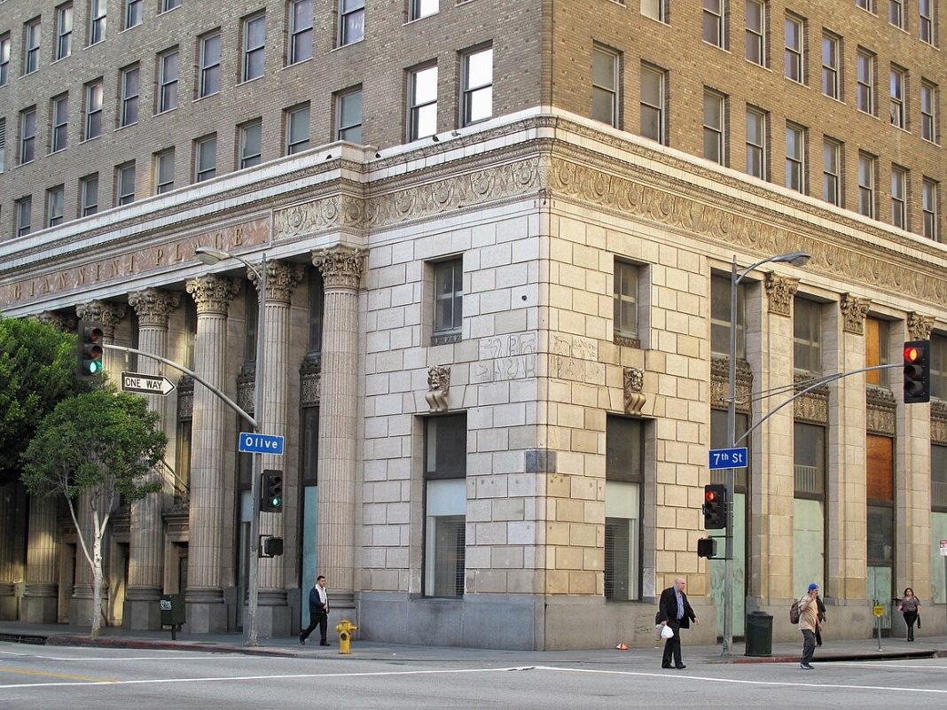 A historic building with ornate columns at the corner of Olive and 7th streets. People walk by, and traffic lights control the intersection.